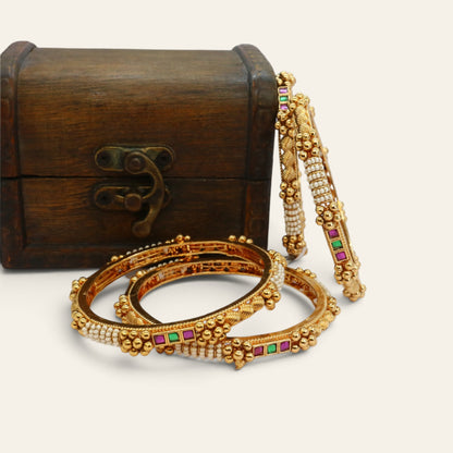 Close-up of four slim gold-tone bangles with rows of pearls and square red and green semi-precious stones