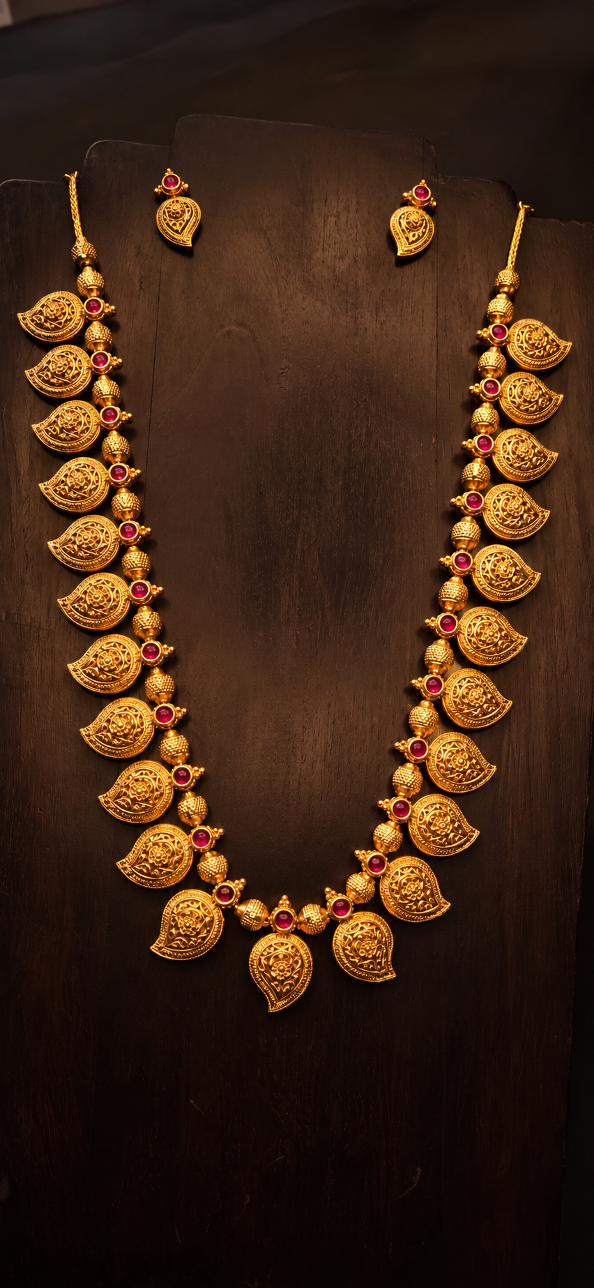 Koyri gold-plated necklace with mango-shaped paisley motifs, textured beads and red accents displayed on dark wood with coordinating earrings above